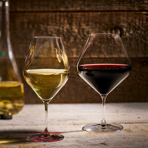 Two Caskata Library White Wine Glasses from a set of 4 sit on a wooden surface, one filled with white wine and the other with red; a Library Collection wine bottle is partially visible in the background.