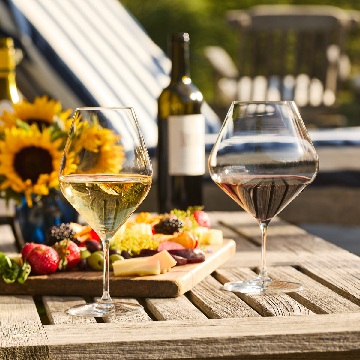Two Caskata Library White Wine Glasses, one with red and one with white wine, sit on a wooden table next to cheese, fruit, a Library Collection wine bottle, and sunflowers in the background.