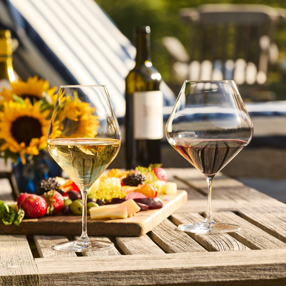 Two Caskata Library White Wine Glasses, one with red and one with white wine, sit on a wooden table next to cheese, fruit, a Library Collection wine bottle, and sunflowers in the background.