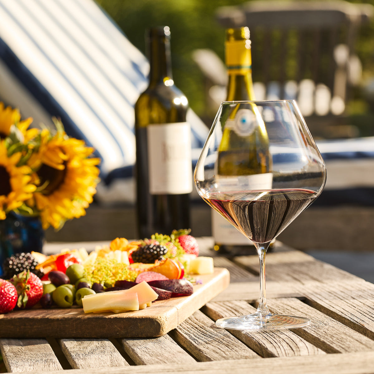 A glass of red wine in a Caskata Library Red Wine Glass from the Set of 4 rests on a wooden table beside a platter of cheeses, fruits, and meats, with two wine bottles and sunflowers in the background.