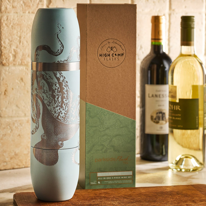 A Caskata x High Camp Light Blue Lucy Parkside Flask with an octopus design stands next to its box on a kitchen counter, with bottles of red and white wine and magnetic tumblers in the background.