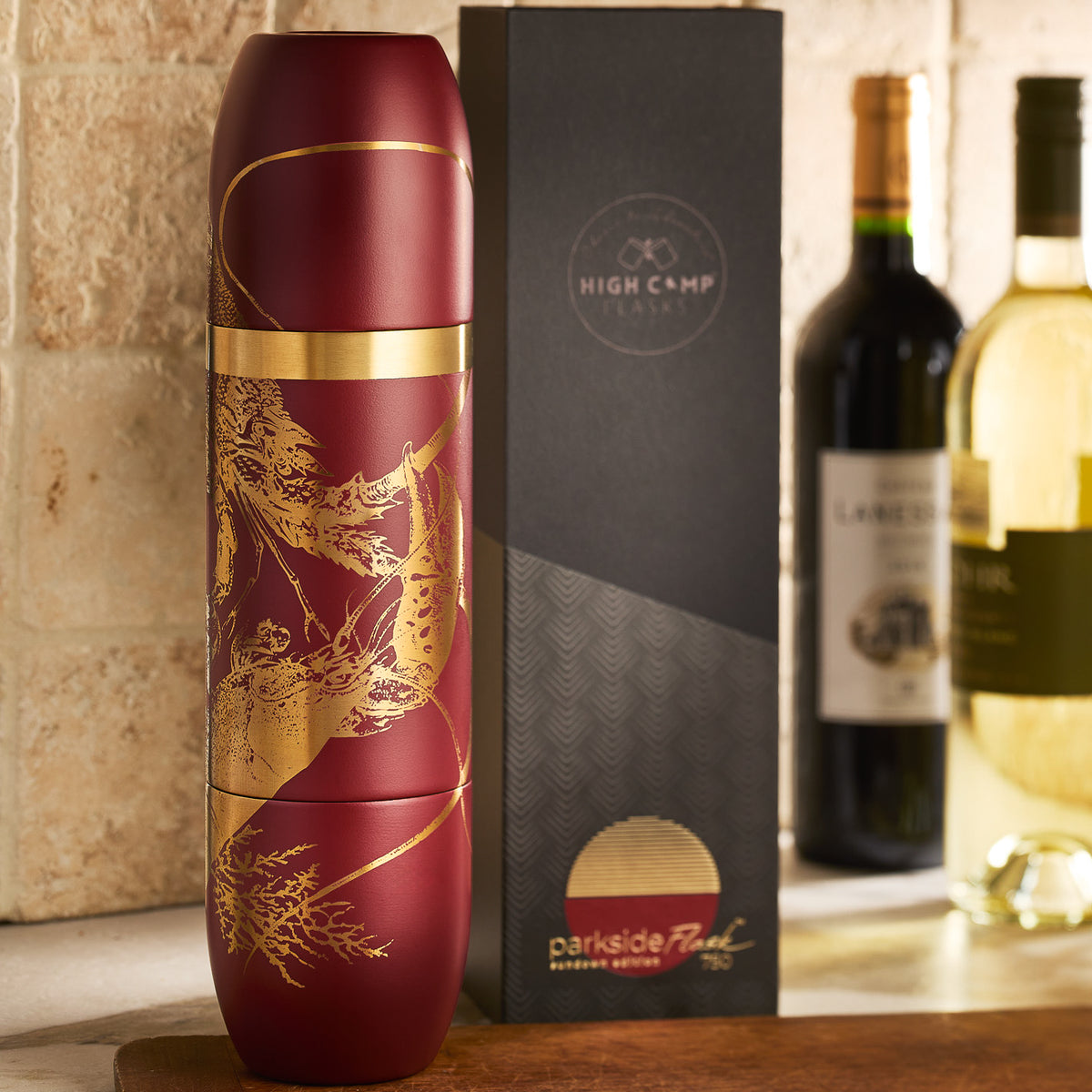 A Caskata x High Camp Red Lobster Parkside Flask by Caskata sits next to its box on a countertop, with two wine bottles in the background.