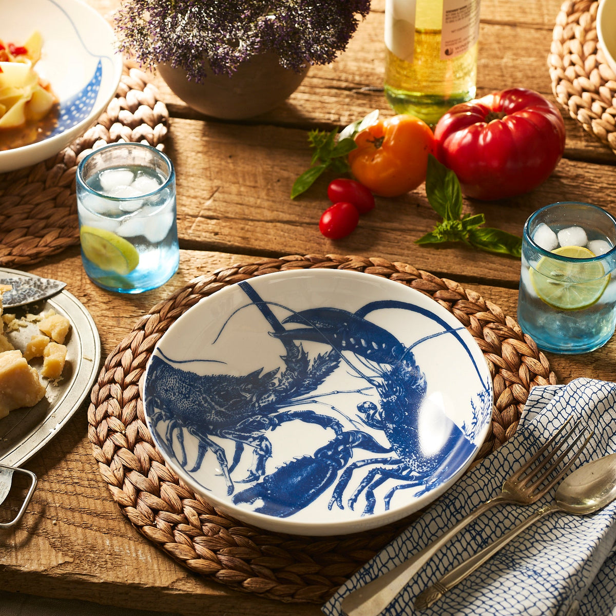 A table set for a meal features the Caskata Lobster Entrée Bowl, adorned with blue lobster illustrations. The stylish and dishwasher-safe porcelain bowl sits among woven placemats, lemon water glasses, bread, tomatoes, and white wine.