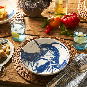 A table set for a meal features the Caskata Lobster Entrée Bowl, adorned with blue lobster illustrations. The stylish and dishwasher-safe porcelain bowl sits among woven placemats, lemon water glasses, bread, tomatoes, and white wine.