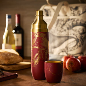 The Caskata x High Camp Red Lobster Parkside Flask by Caskata sits on a wooden table with apples, bread, wine bottles, and a patterned tote bag in the background.
