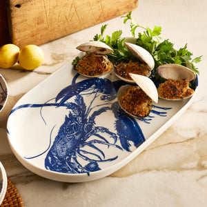 The Caskata Lobster Large Coupe Oval Platter displays baked stuffed clams, fresh parsley, and clam shells, with lemons in the background on marble—a stylish entertaining serveware piece.