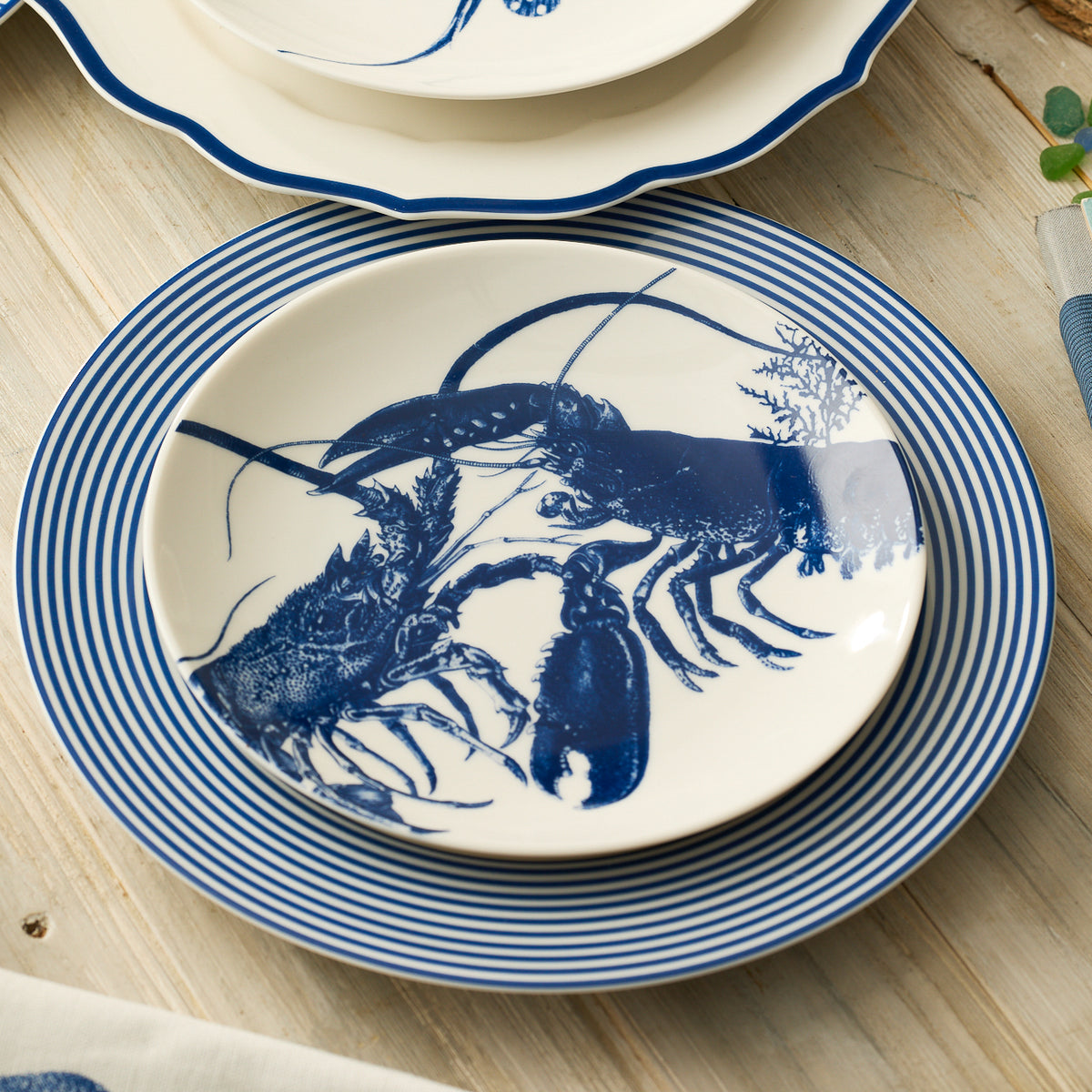 The Caskata Lobster Coupe Salad Plate, decorated with a blue lobster, tops two striped plates to bring coastal charm to a light wood table.