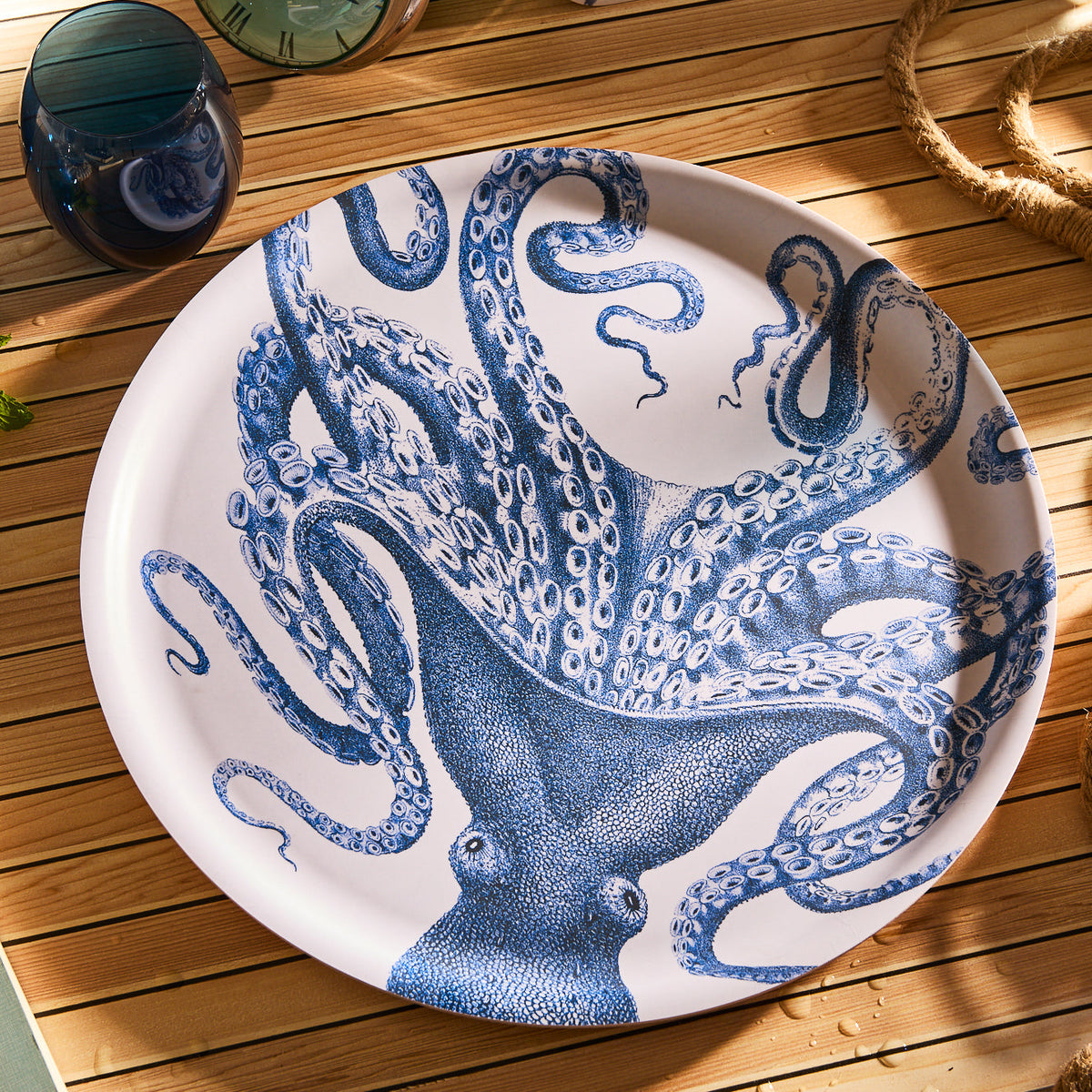 The Lucy Large Round Birchwood Tray by Caskata features a detailed blue octopus illustration. It's displayed on a wooden table with a blue glass, rope, and greenery in the background. Handcrafted in Finland, this tray seamlessly blends artistry with tradition for a striking display.