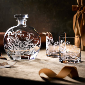 The Lucy by the Fireside Bundle by Caskata features a crystal decanter and two octopus-etched glasses, arranged on fabric with a brown ribbon and soft lighting for an impressive whiskey gift set.