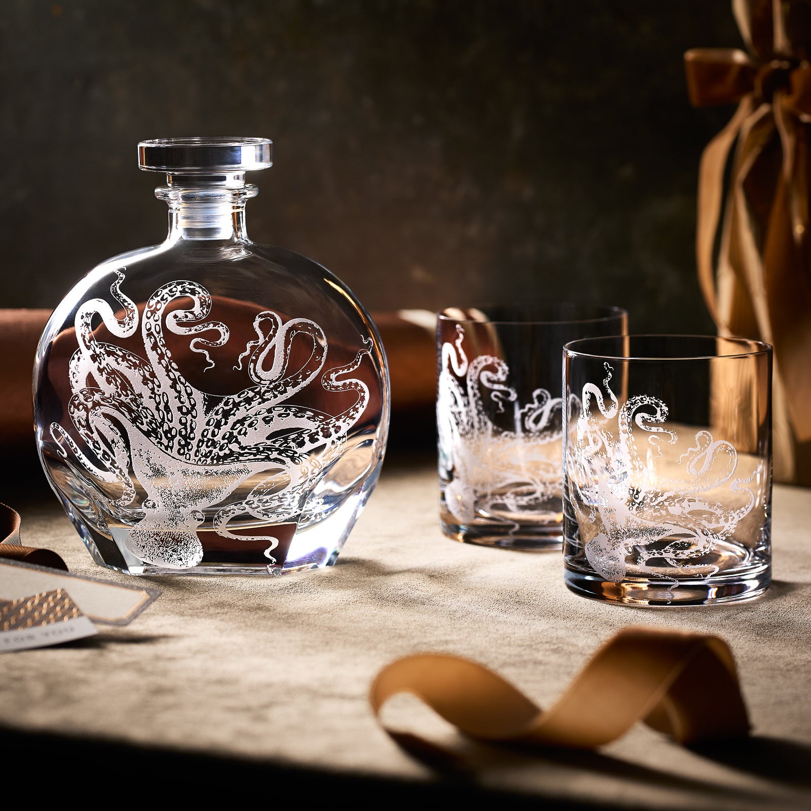 The "Lucy by the Fireside" Bundle from Caskata includes a lead-free crystal decanter and two rocks glasses with octopus etching, arranged on beige fabric beside a brown ribbon.