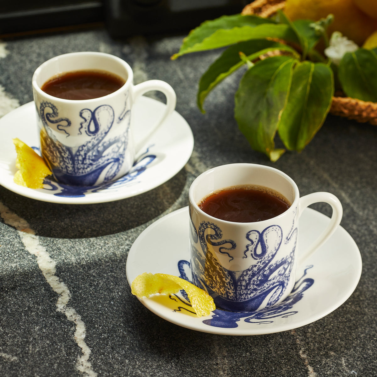 A Lucy Espresso Cup &amp; Saucer Set/2 of tea on a contemporary patterned table next to a slice of lemon.