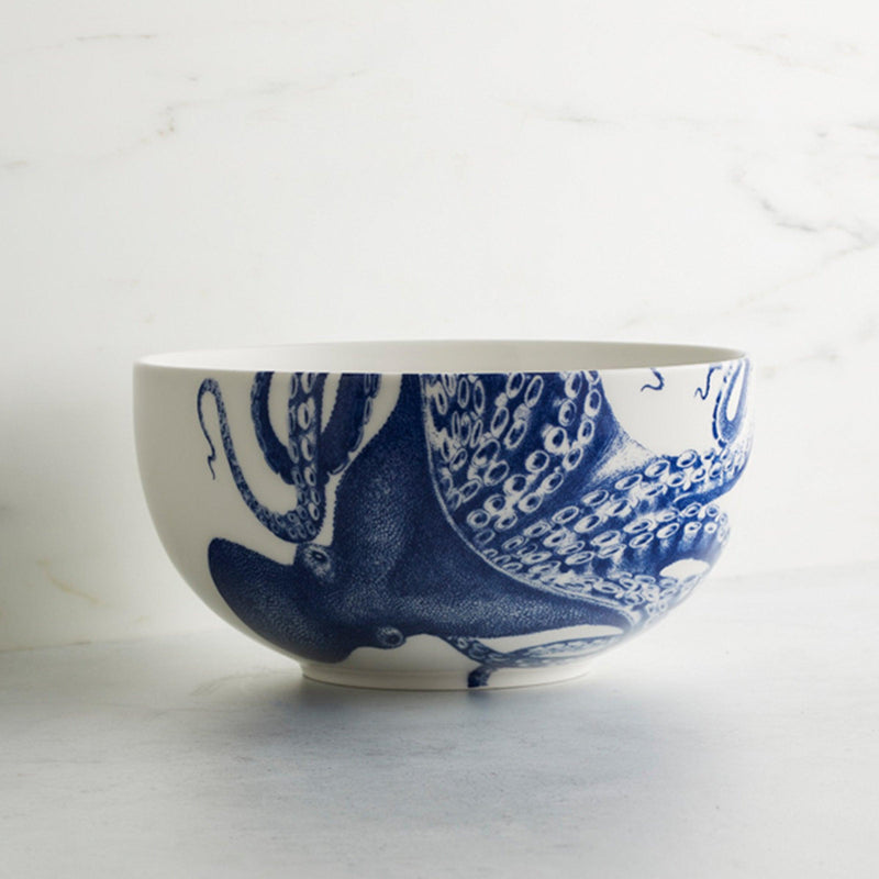 A premium porcelain serving bowl, known as the Lucy Large Round Serving Bowl by Caskata Artisanal Home, features a blue octopus design with tentacles wrapping around the exterior. It is placed on a light surface with a white background, embodying elegant blue and white tableware.