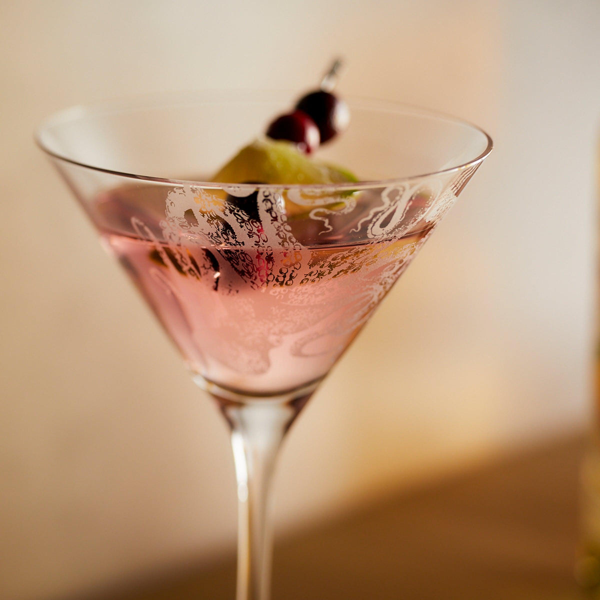 A close-up of a pink cocktail in a Caskata Artisanal Home Lucy Martini Glasses garnished with a lime twist and two red cherries on a pick.