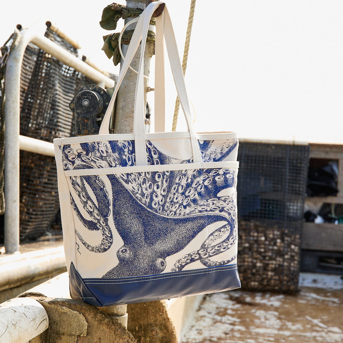 The Caskata Lucy the Octopus Caskata x Steele Everyday Tote Bag, featuring blue handles and an octopus design, hangs on a metal railing outdoors near fishing equipment.