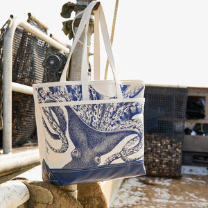 The Caskata Lucy the Octopus Caskata x Steele Everyday Tote Bag, featuring blue handles and an octopus design, hangs on a metal railing outdoors near fishing equipment.