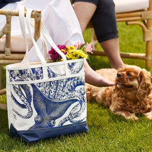 The Caskata Lucy the Octopus Everyday Tote Bag features a blue octopus print and sits on grass by a chair, filled with flowers. A brown dog rests nearby with a person's legs visible—an ideal handcrafted tote for stylish outings.