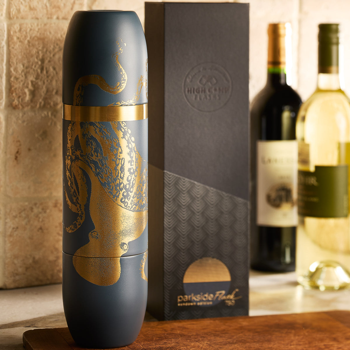 The Caskata x High Camp Dark Blue Lucy Parkside Flask, featuring a matte finish and gold octopus design, stands before its box beside wine bottles on a kitchen counter—an eye-catching piece from Caskata.