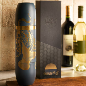 The Caskata x High Camp Dark Blue Lucy Parkside Flask, featuring a matte finish and gold octopus design, stands before its box beside wine bottles on a kitchen counter—an eye-catching piece from Caskata.
