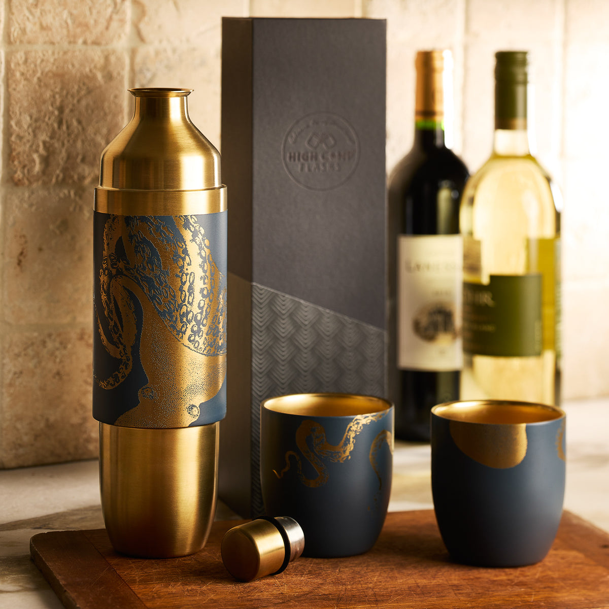 A gold and navy cocktail shaker set with two matching cups sits on a counter; behind it are two bottles of wine, a black gift box, and the Caskata x High Camp Dark Blue Lucy Parkside Flask by Caskata.