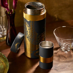 A metallic black and gold thermos with an open lid, matching cup, etched glass, plate, floral vase, and a sleek cocktail shaker from Caskata's Retro Beach Bar Bundle sit on a wooden surface.