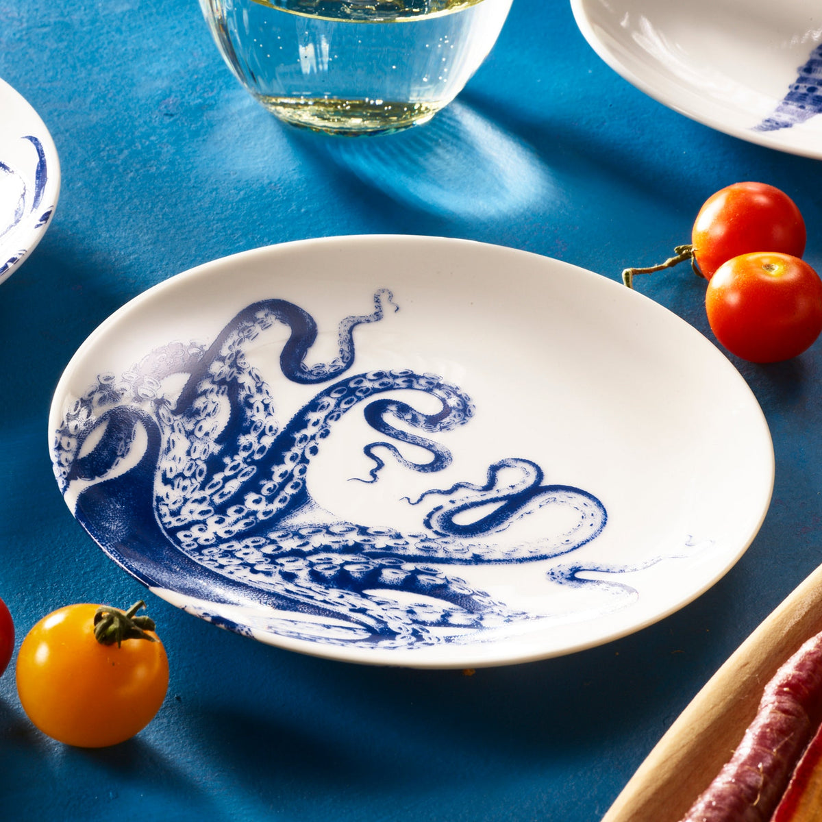 A white plate with an octopus pattern design, positioned on a blue surface with various small tomatoes and an out-of-focus glass in the background, showcases the style of Lucy Small Plates from Caskata Artisanal Home.