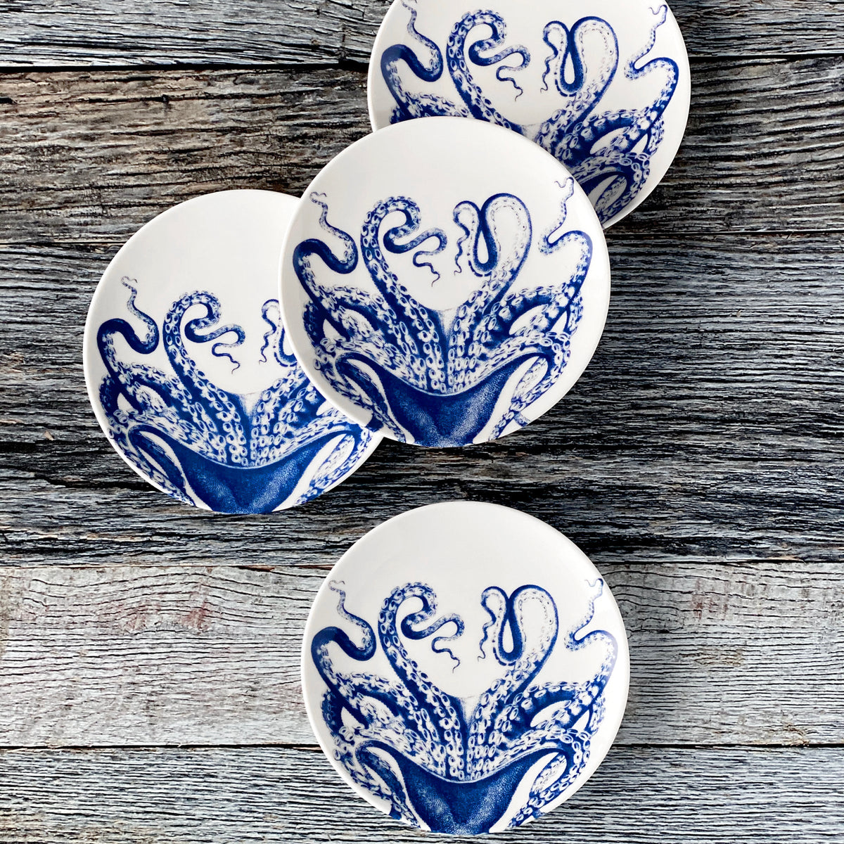 Four heirloom-quality Lucy Small Plates by Caskata Artisanal Home, featuring intricate blue octopus designs, are arranged beautifully on a textured wooden surface.