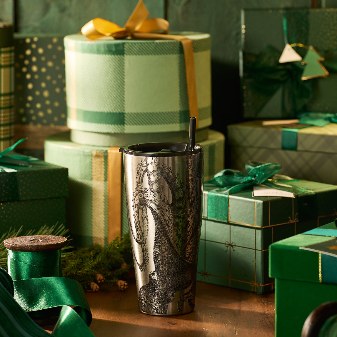 The Caskata Lucy Chrome Stainless Steel Insulated Tumbler with an octopus design and straw is on a wooden table, surrounded by green and gold wrapped gift boxes.