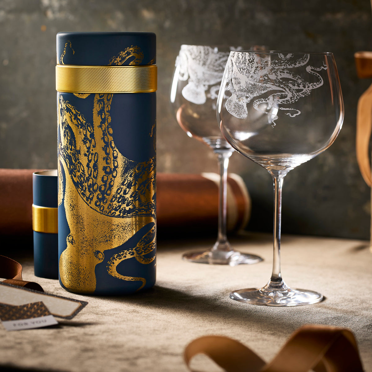The Pleased as Punch Bar Bundle by Caskata features a navy and gold octopus cylindrical container and two gin and tonic glasses with octopus motifs—a chic barware set for your home.