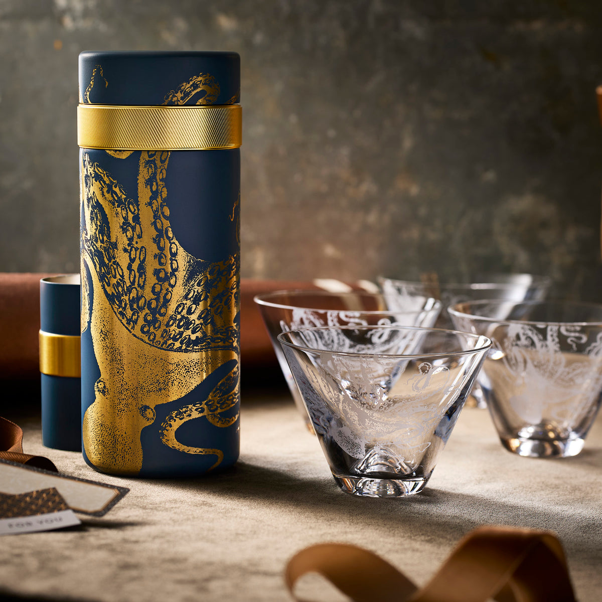 The Retro Beach Bar Bundle by Caskata features a navy blue cylindrical cocktail shaker with a gold octopus design, set next to three etched glass tumblers on a stone surface.