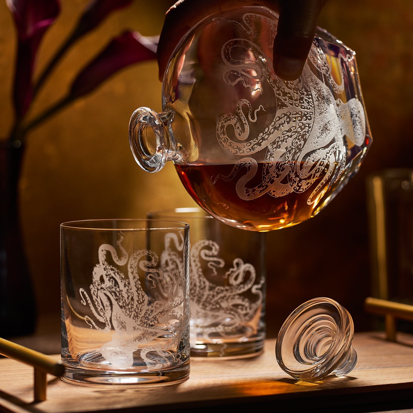 A hand pours amber liquid from the Caskata Artisanal Home Lucy Decanter, its sand-etched octopus design reflected on matching glasses, with the stopper resting nearby.