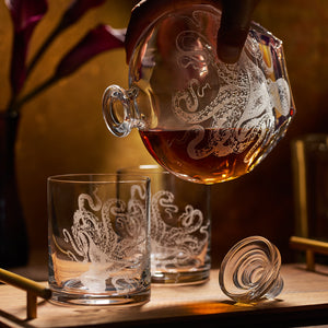 A hand pours amber liquid from the Caskata Artisanal Home Lucy Decanter, its sand-etched octopus design reflected on matching glasses, with the stopper resting nearby.