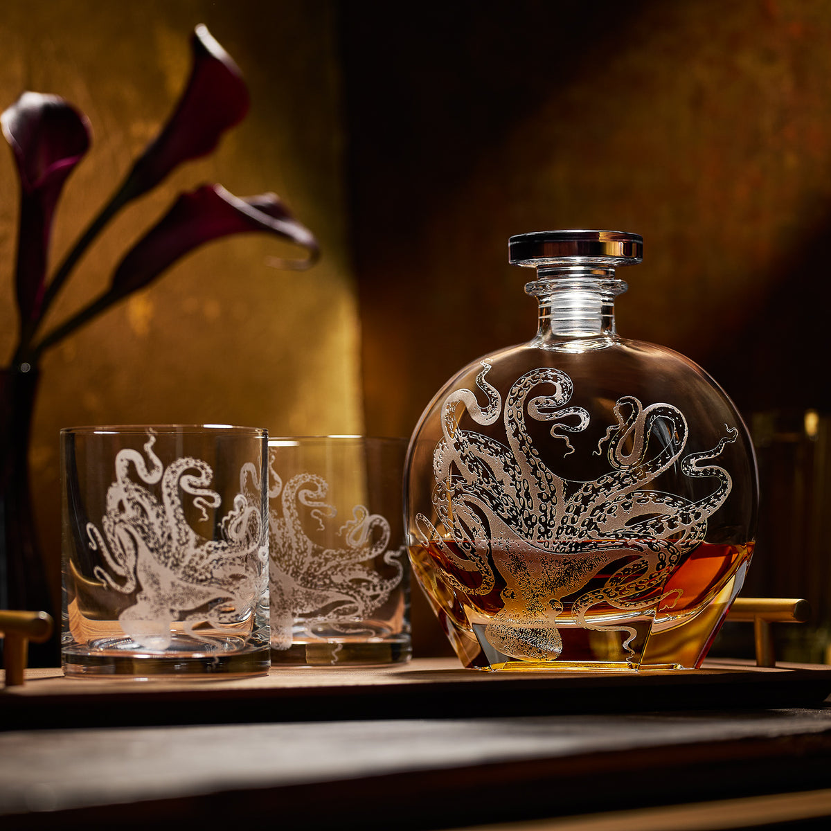 The Lucy Decanter by Caskata Artisanal Home and two matching tumblers, each featuring a sand-etched octopus design, rest on a tray. Purple calla lilies form a striking backdrop to this unique crystal glassware set.