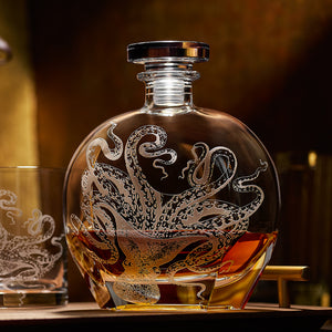 The Lucy Decanter by Caskata Artisanal Home, expertly sand-etched on crystal glass and partially filled with brown liquor, sits beside a matching glass.