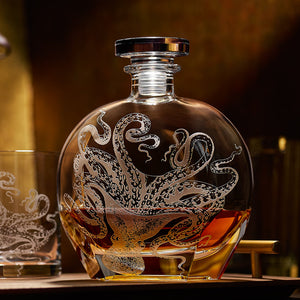 The Lucy Decanter by Caskata Artisanal Home, expertly sand-etched on crystal glass and partially filled with brown liquor, sits beside a matching glass.