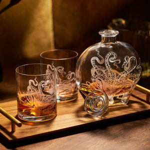The Lucy Decanter by Caskata Artisanal Home, etched with an octopus, and two crystal glasses filled with amber liquid sit on a wooden tray with a gold handle under warm lighting.
