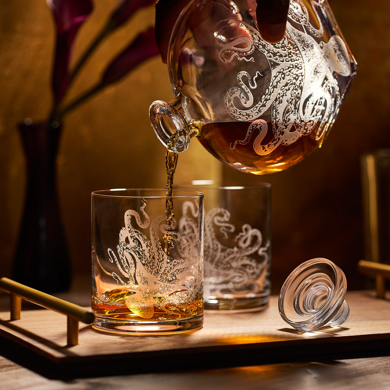 A hand pours amber liquid from the sand-etched Lucy Decanter by Caskata Artisanal Home into a matching crystal glass on a wooden tray, with the lid and another glass nearby.