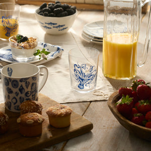 A breakfast table with orange juice in a Caskata Lucy Blues Short Drinking Glass, a plate of strawberries, muffins, yogurt parfaits, and blackberries on a wooden surface.