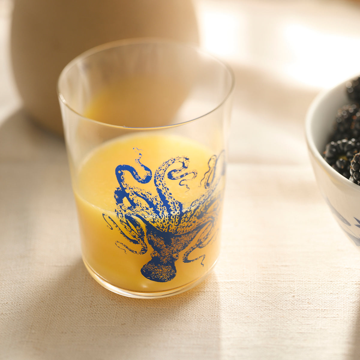 The Caskata Lucy Blues Short Drinking Glass, adorned with a blue octopus, sits on a table filled with orange juice next to a bowl of blackberries.