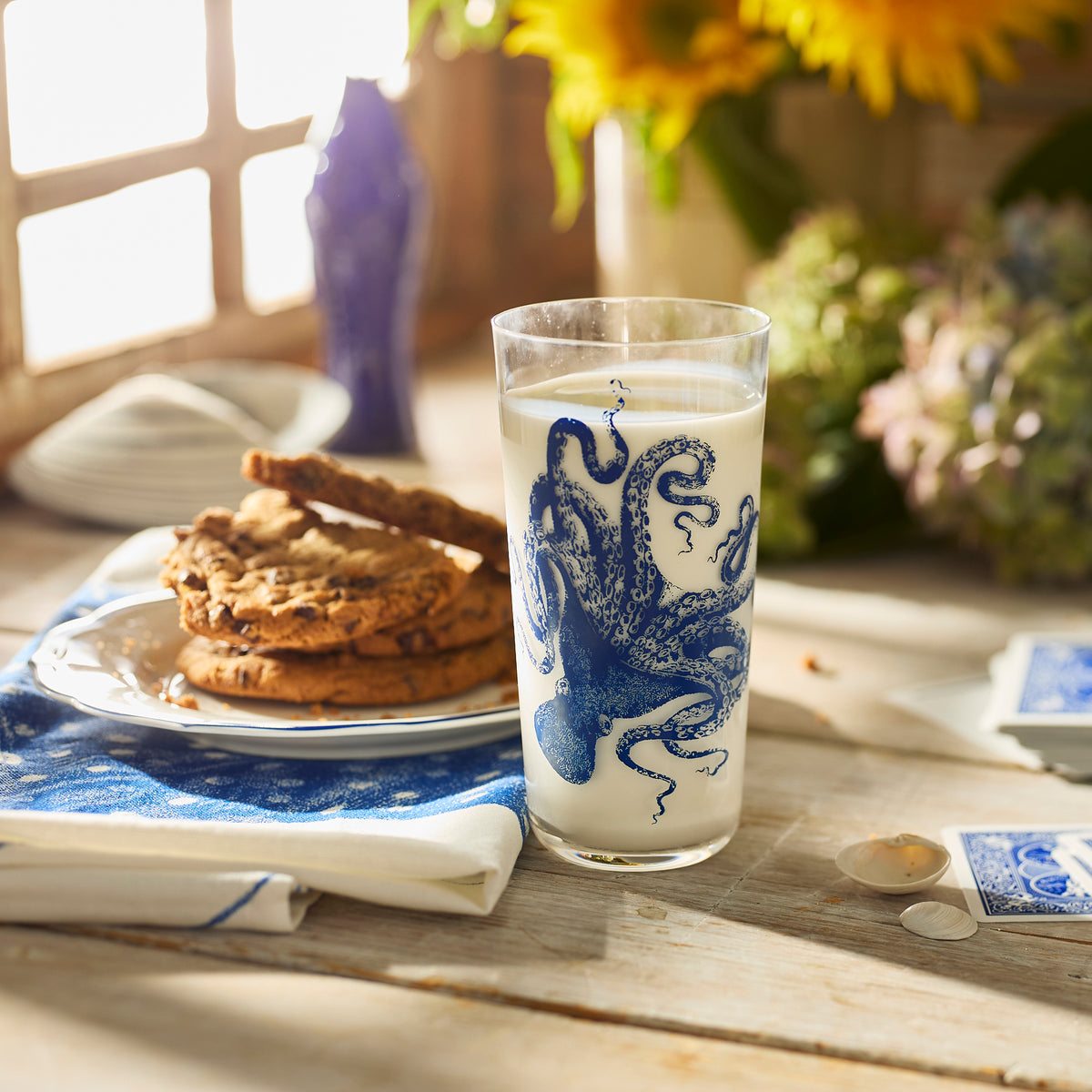 A glass of milk in a Caskata Lucy Blues Tall Drinking Glass featuring an octopus sits by cookies on a wooden table, with flowers and sunlight in the background.