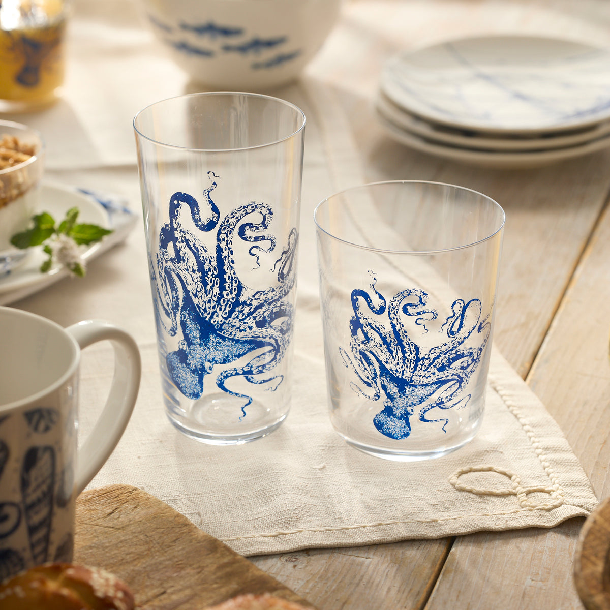 Two Caskata Lucy Blues Tall Drinking Glasses, crafted from lead-free crystal and featuring blue octopus designs, rest on a table with a beige napkin, surrounded by plates and other tableware.