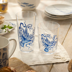 Two Caskata Lucy Blues Tall Drinking Glasses, crafted from lead-free crystal and featuring blue octopus designs, rest on a table with a beige napkin, surrounded by plates and other tableware.