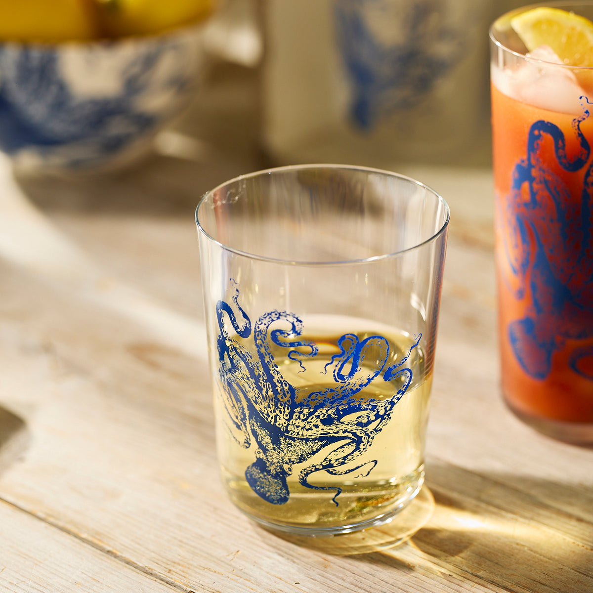 A Lucy Blues Short Drinking Glass by Caskata holds a light drink beside another with a reddish beverage and ice, both highlighting the elegance of Caskata luxury tumblers crafted from lead-free crystal.