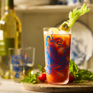 A Bloody Mary garnished with celery, lemon, and olives in a lead-free crystal Caskata Lucy Blues Tall Drinking Glass, set on a wooden board with tomatoes, basil, and a wine bottle in the background.