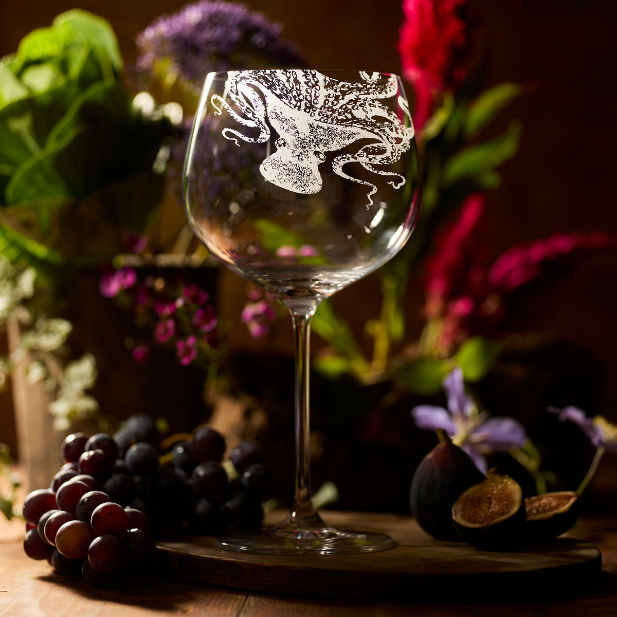 The Caskata Lucy Gin &amp; Tonic Glass features a lead-free crystal design with an octopus motif, shown on a wooden surface with grapes, figs, and flowers in the background.