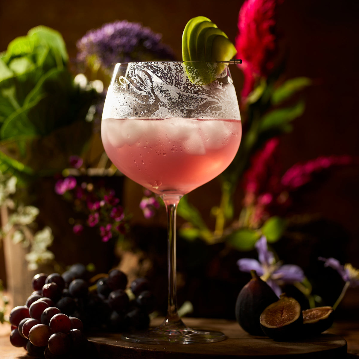 A large pink cocktail with ice and a lime garnish is served in a Caskata Lucy Gin & Tonic Glass, resting on a rustic surface with grapes, figs, and flowers in the background.
