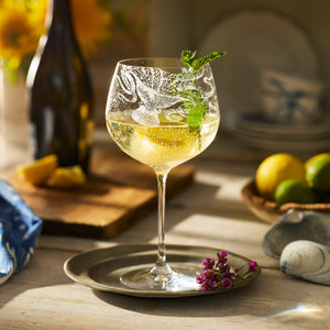 A Caskata Lucy Gin & Tonic Glass, etched and stemmed, holds a sparkling yellow drink with mint on a metal plate, surrounded by lemons, limes, flowers, and a wine bottle on a sunlit table.