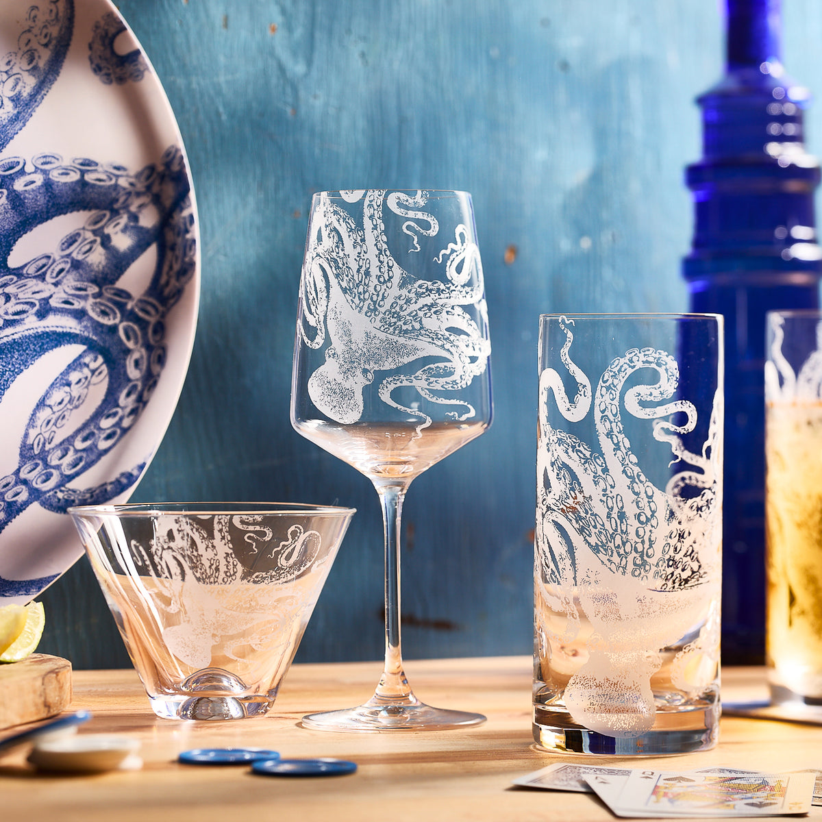 Glassware and a plate with octopus designs are on a wooden surface. A dark blue bottle is in the background, with Lucy Highball Glasses by Caskata Artisanal Home sparkling nearby. Cards and a lime slice complete the scene.