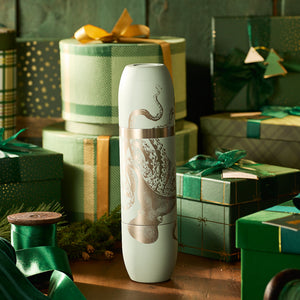 A white ceramic vase with an octopus design sits on a wooden table, surrounded by green and gold gift boxes, festive decor, and the Caskata x High Camp Light Blue Lucy Parkside Flask from Caskata, ready for celebratory toasts.