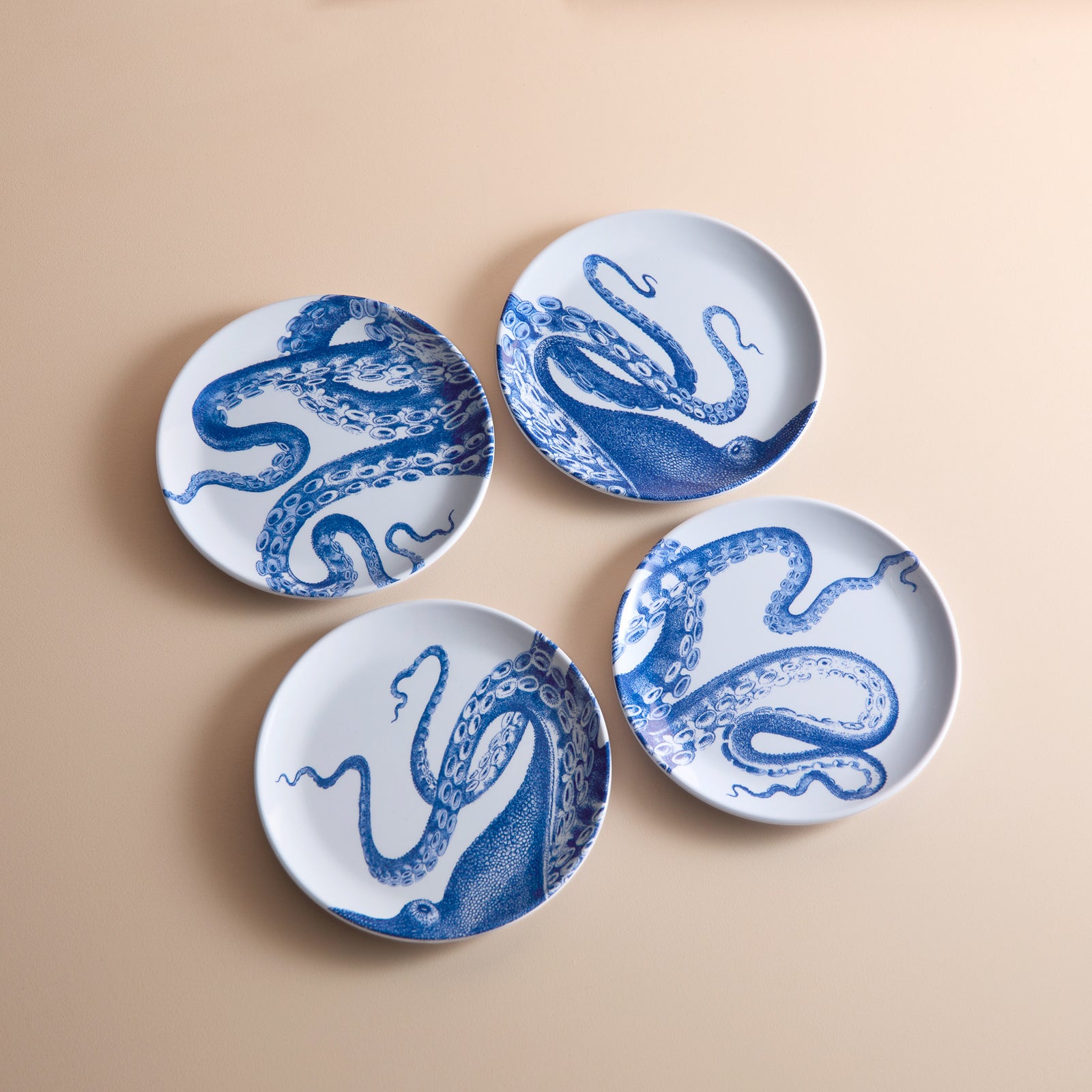 A set of four Lucy Melamine Small Plates by Caskata, featuring blue octopus designs, is displayed on a beige surface, highlighting their unbreakable style for any occasion.