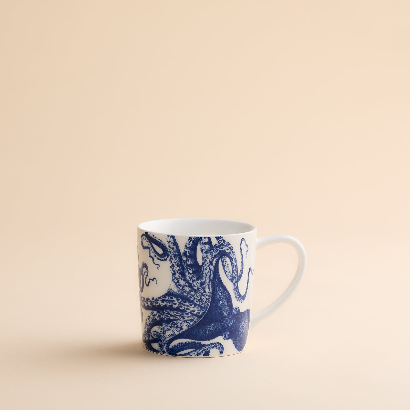 The Lucy Mug by Caskata Artisanal Home features a charming blue octopus illustration on white porcelain, displayed against a simple beige surface and background—perfect for bringing a coastal vibe to your morning routine.
